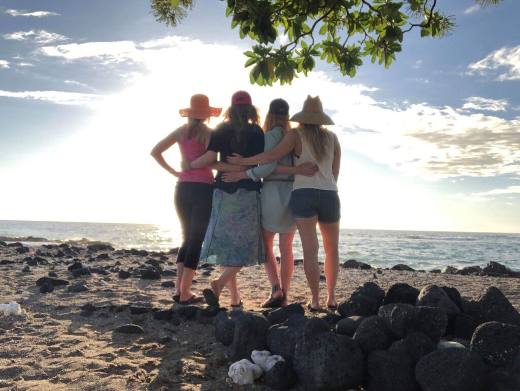 A group of woman standing on a beach with their backs to the camera, embracing and looking out at the ocean.