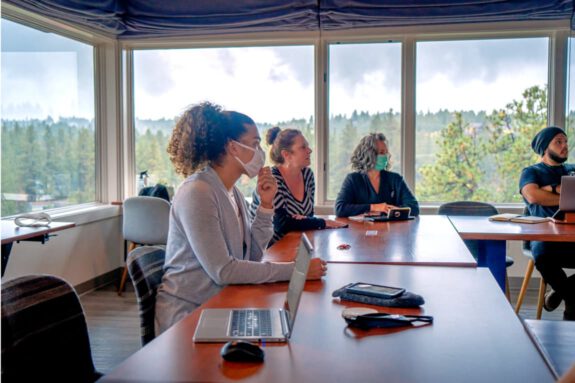 A group of Haven members having a meeting and watching a presentation in The Library with windows overlooking the Deschutes River.