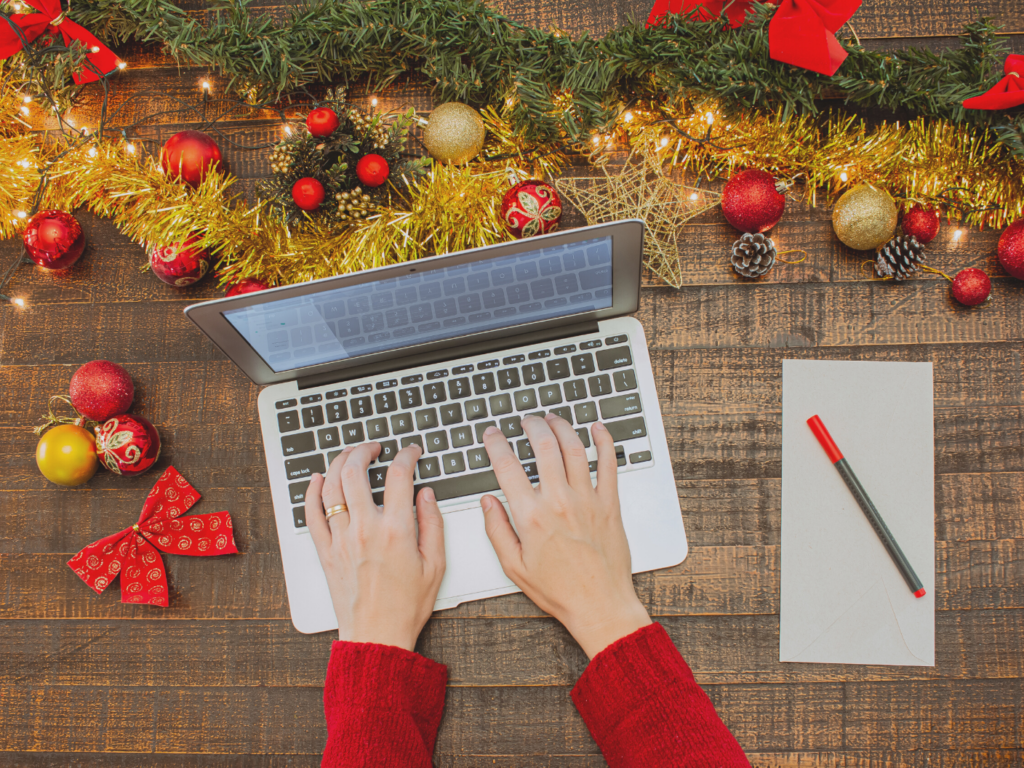 Overhead view of woman working on laptop surrounded by garland and christmas ornaments making a list of gift ideas for remote workers