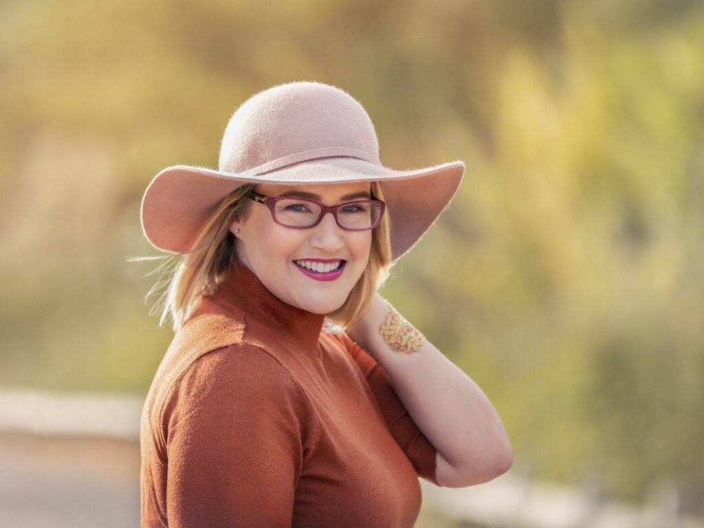 rachelle indra poses with a rust colored long sleeve shirt, tan hat, glasses, and holding her hand behind her hat