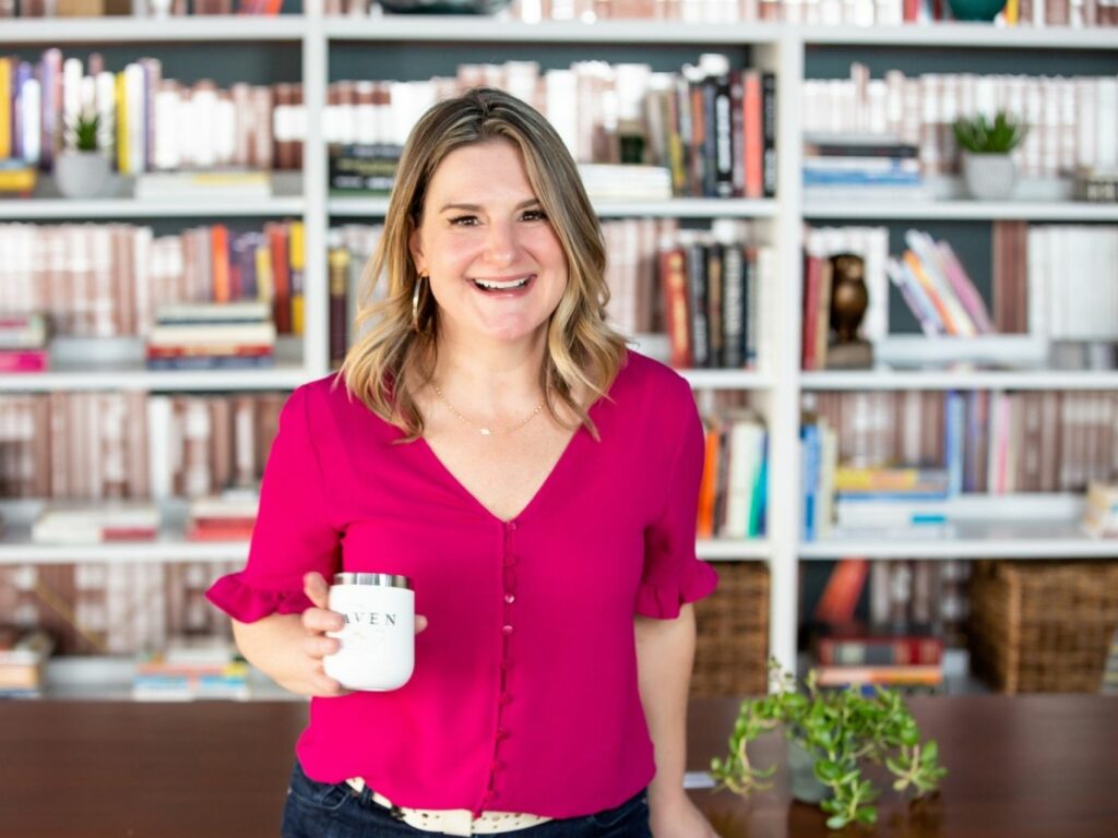 christy lawrence, woman in pink shirt and blonde hair, holds a coffee cup in front of a bookshelf and smiles