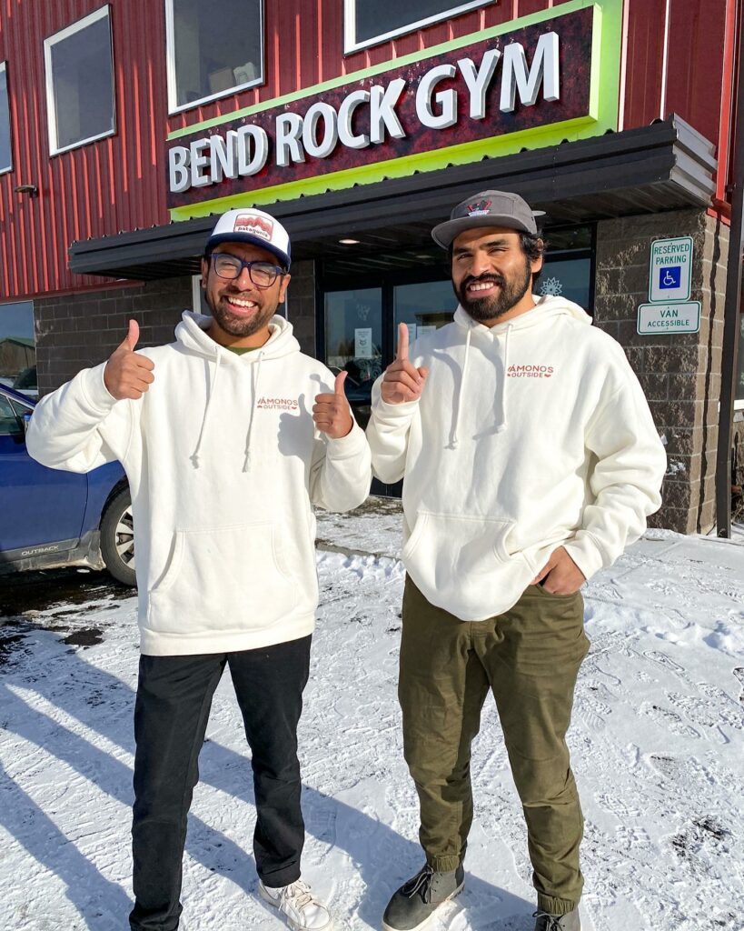 Two men wearing white sweatshirts and ball caps stand outside Bend Rock Gym building while giving thumbs pu