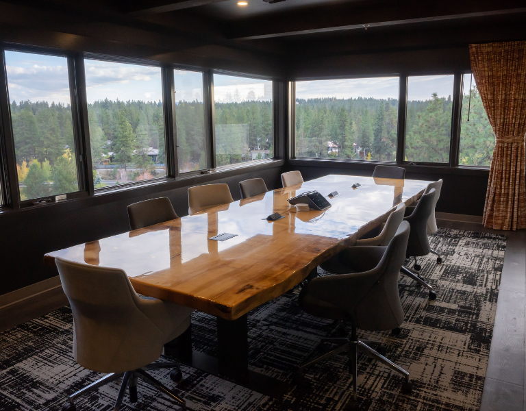 long wooden table lined with suede chairs sits within the hinterland meeting room at the haven coworking, with large windows looking over the deschutes river.