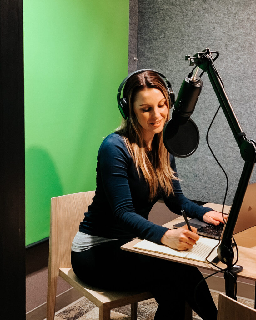 woman wearing headphones speaks into a podcasting microphone with a green screen in the background.